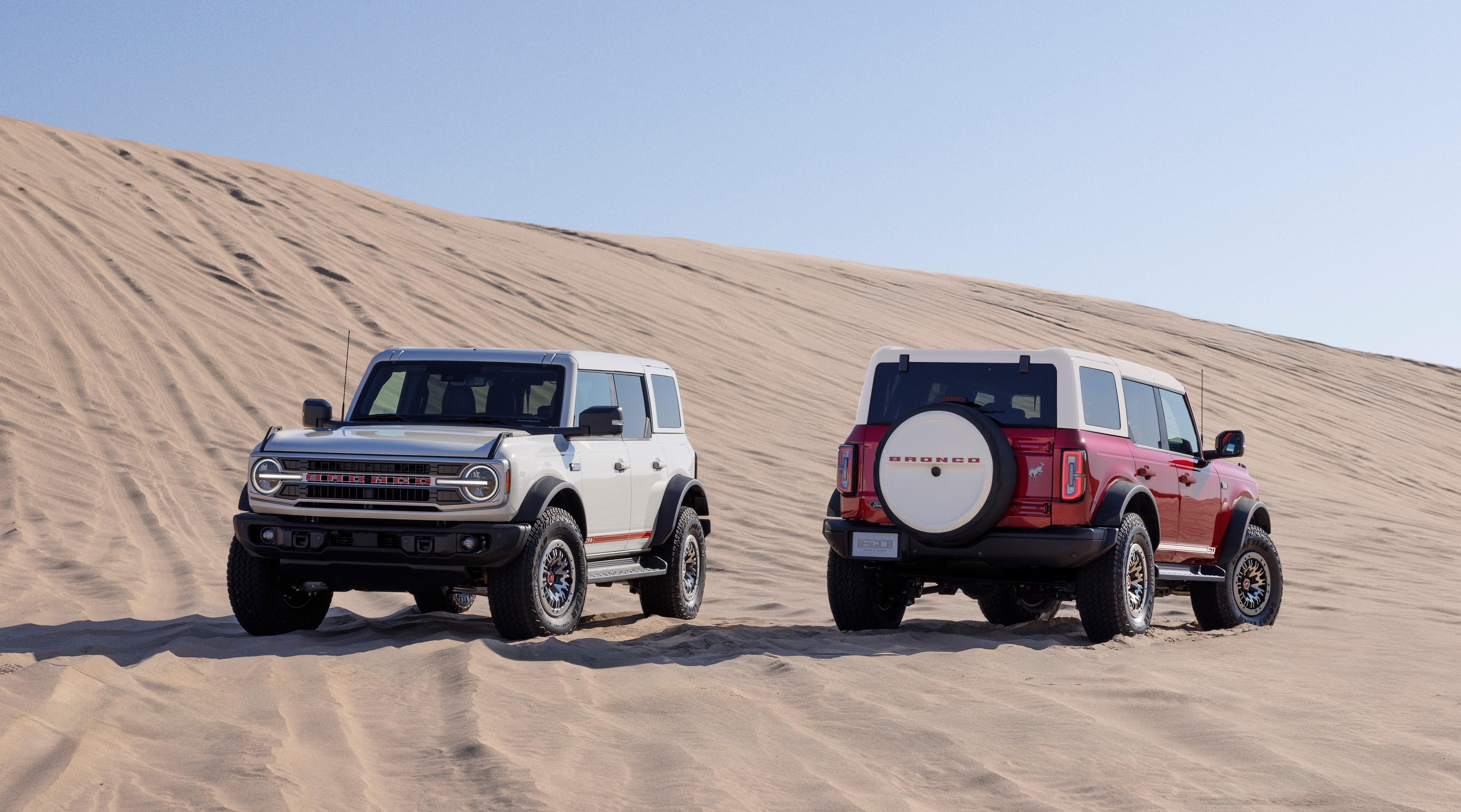 2026 Ford Bronco 60th Anniversary Package shown on two Broncos, one white with red accents and one red with white accents. Vehicles are parked on a sandy hill with a blue sky overhead.
